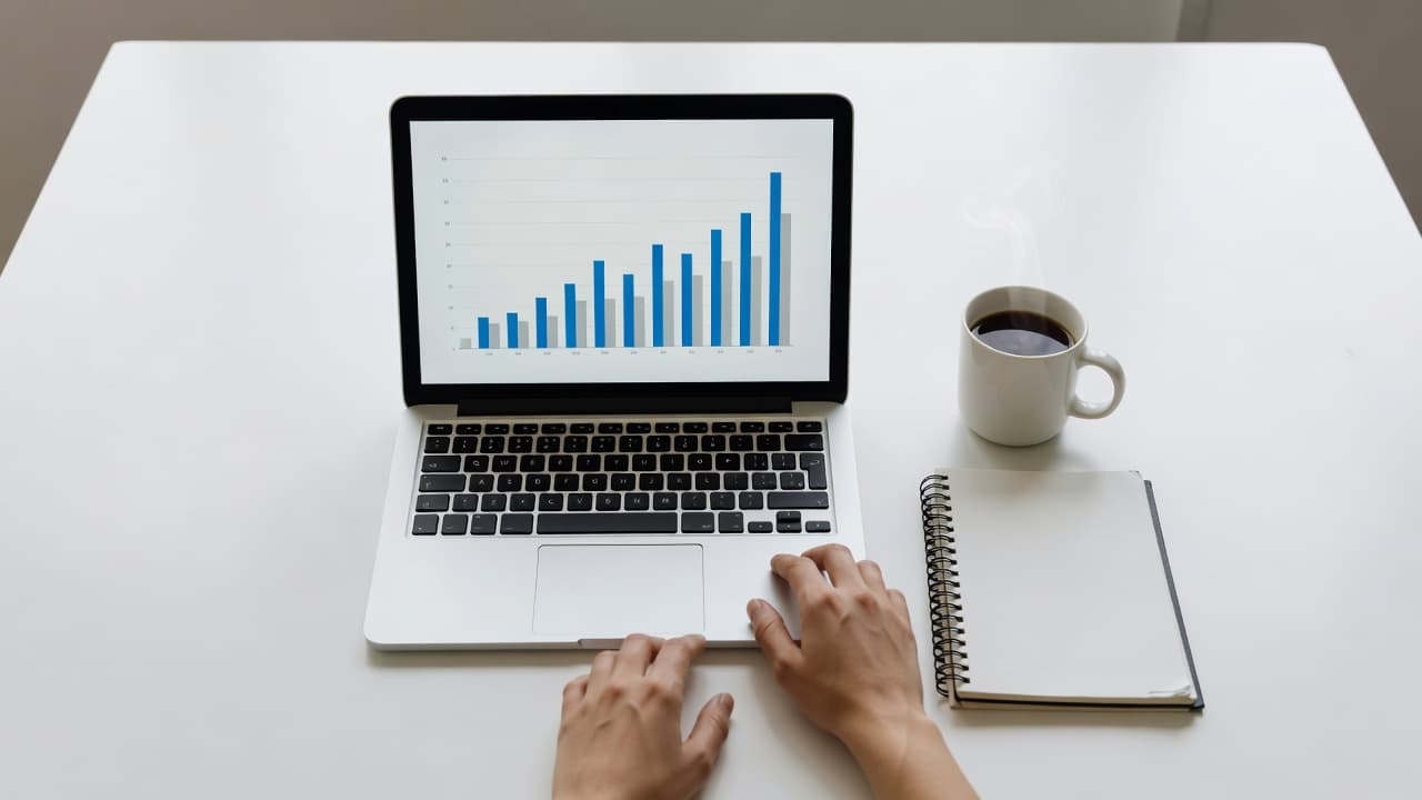 A close view shows a person's hands resting on a clean desk beside a laptop screen displaying rising bar charts. A ceramic coffee mug and spiral notebook sit nearby under bright light.